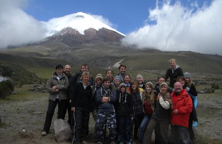 Eine Gruppe von Menschen posiert fröhlich vor dem schneebedeckten Cotopaxi-Vulkan unter einem klaren blauen Himmel.