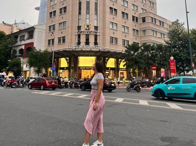Eine Frau in einem gestreiften Oberteil und einem rosa Rock steht auf der Straße vor einem eleganten Gebäude in Saigon.