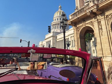 Im Vordergrund sind fröhliche Passagiere in einem pinken Cabrio zu sehen, während sie an historischen Gebäuden von Havanna vorbeifahren.