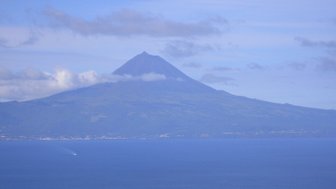 Ein majestätischer Vulkan erhebt sich über das ruhige blaue Wasser, umgeben von sanften Wolken und einem klaren Himmel.