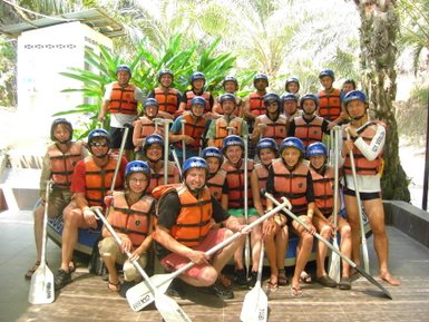 Eine Gruppe von 20 Personen in Schwimmwesten und Helmen posiert fröhlich vor einer tropischen Kulisse.