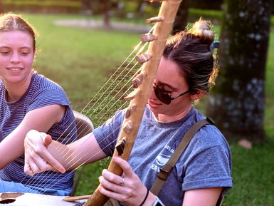 Eine Frau spielt konzentriert auf einer traditionellen Harfe, während eine andere Person interessiert zuschaut.