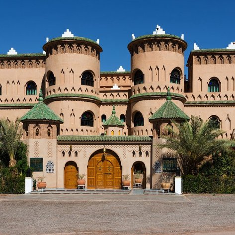 Ein beeindruckendes, maurisches Gebäude mit grünen Dächern und kunstvollen Fenstern, umgeben von Palmen und Pflanzen.