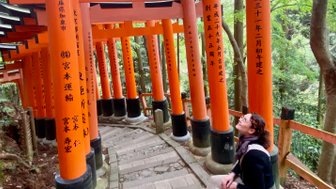 Eine Person sitzt auf einer Steinbank und blickt lächelnd zu den leuchtend orangefarbenen Torii-Toren auf einem schmalen Weg.