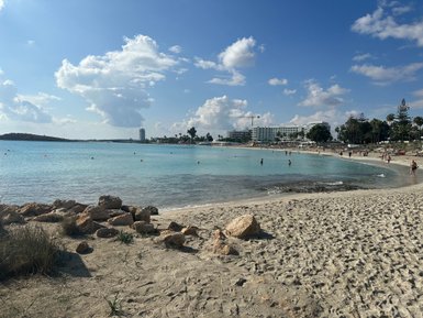 Ein sonniger Strand mit feinem Sand, klarem Wasser und Menschen, die sich am Ufer entspannen und schwimmen.