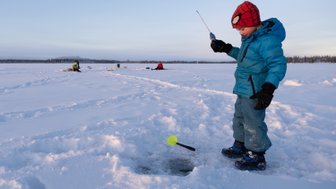 Ein Junge in blauer Winterkleidung steht auf dem Schnee und angelt durch ein Loch im Eis, während andere im Hintergrund fischen.