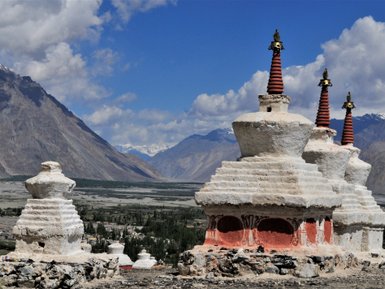 Drei beeindruckende Stupas mit roten Spitzen stehen vor einer majestätischen Berglandschaft unter einem blauen Himmel.