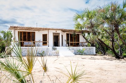 Ein modernes, weißes Haus mit Holzveranda und Palmen, umgeben von sandigem Boden und üppiger Vegetation.