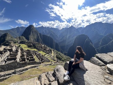 Eine Person sitzt auf einem großen Stein und blickt auf die beeindruckenden Ruinen von Machu Picchu und die umliegenden Berge.