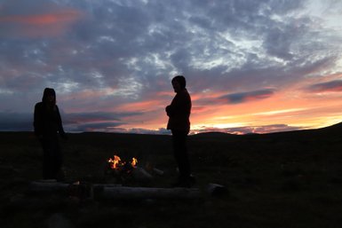 Zwei Personen stehen am Lagerfeuer, umgeben von einer malerischen Landschaft bei Sonnenuntergang.
