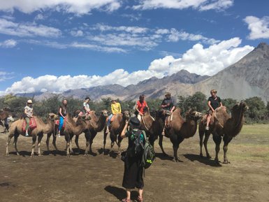 Eine Gruppe von Menschen reitet auf Kamelen in einer malerischen Landschaft mit Bergen und blauem Himmel.