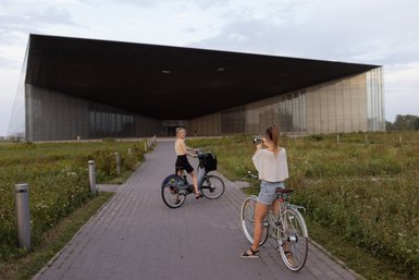 Zwei Frauen auf Fahrrädern stehen auf einem Weg, während eine von ihnen die andere fotografiert, vor einem modernen Gebäude.