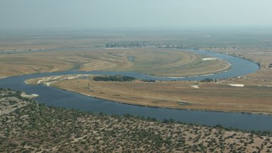 Eine weite Landschaft mit einem geschwungenen Fluss, umgeben von goldenen Wiesen und vereinzelten Bäumen im Hintergrund.