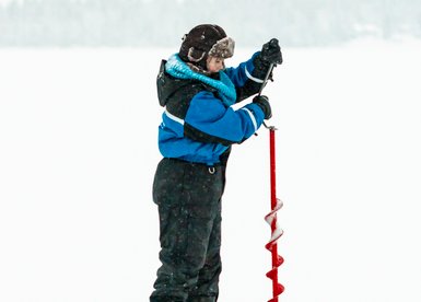 Ein Kind steht auf einer schneebedeckten Fläche und bohrt mit einer roten Eisbohrmaschine ein Loch ins Eis.