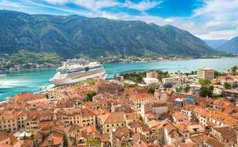 Eine atemberaubende Panoramaansicht von Kotor mit einem großen Kreuzfahrtschiff im Hafen und den majestätischen Bergen im Hintergrund.