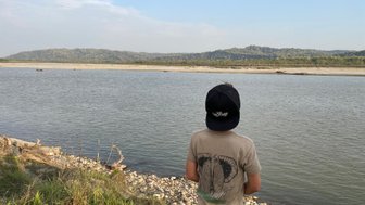 Ein Junge steht am Ufer eines ruhigen Flusses und blickt auf die sanften Hügel im Hintergrund unter einem klaren Himmel.