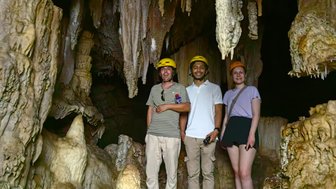 Drei Personen stehen in einer Höhle, umgeben von beeindruckenden Stalaktiten und Stalagmiten, während sie Helme tragen.
