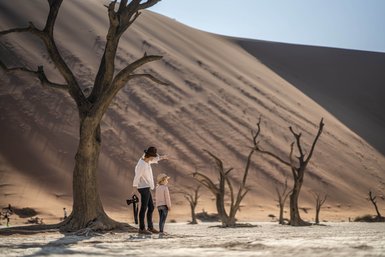 Eine Mutter zeigt mit ihrer Hand auf die beeindruckenden Dünen von Deadvlei, während ihr Kind aufmerksam zusieht.
