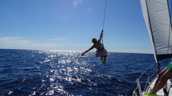 Eine Person schwingt fröhlich über dem glitzernden Wasser, während sie an einem Seil von einem Segelboot hängt.