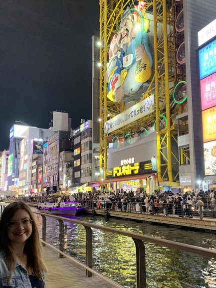 Eine Frau mit Brille lächelt am Ufer des Dotombori-Flusses, umgeben von bunten Neonlichtern und Menschenmengen.
