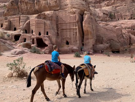 Zwei Kinder reiten auf Eseln in der antiken Stadt Petra, umgeben von beeindruckenden Felsformationen und Höhlen.