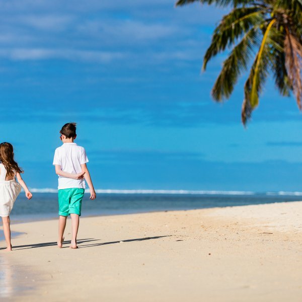 Eine Mutter und zwei Kinder spazieren barfuß am Strand entlang, umgeben von Palmen und einem strahlend blauen Himmel.