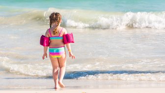 Ein kleines Mädchen mit Zöpfen trägt bunte Schwimmflügel und geht fröhlich zum Wasser am Strand.