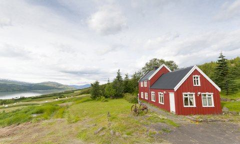 Ein rotes Sommerhaus steht malerisch in einer grünen Landschaft mit Blick auf einen ruhigen See und sanfte Hügel im Hintergrund.