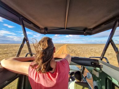 Eine Frau mit langen Haaren lehnt entspannt aus einem Geländewagen und genießt die Aussicht auf die weite Savanne.