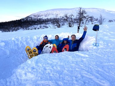 Eine Familie sitzt im Schnee, umgeben von schneebedeckten Bergen, und winkt fröhlich in die Kamera.