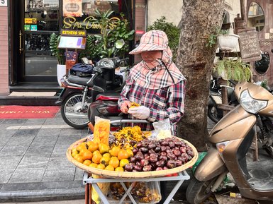 Eine Straßenverkäuferin in Hanoi sortiert frisches Obst und Snacks an ihrem Wagen vor einem Spa.