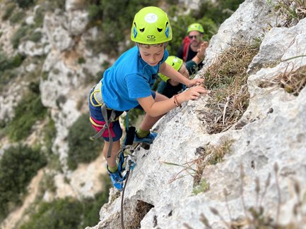 Ein junger Kletterer mit einem grünen Helm erklimmt eine steile Felswand, während ein weiterer Kletterer im Hintergrund zusieht.