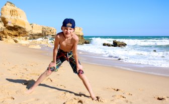 Ein fröhlicher Junge steht am Strand, mit weit gespreizten Beinen und einem breiten Lächeln, während die Wellen sanft an den Strand rollen.