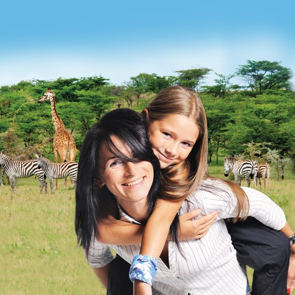 Eine Frau trägt ein Mädchen auf dem Rücken, während im Hintergrund Giraffen und Zebras in einer grünen Landschaft zu sehen sind.