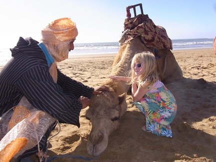 Ein Mann in traditioneller Kleidung streichelt ein Kamel am Strand, während ein kleines Mädchen mit Sonnenbrille daneben steht.
