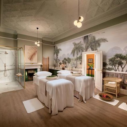 Ein eleganter Spa-Raum mit mehreren Massagetischen, umgeben von beruhigenden Farben und Pflanzenmotiven an der Wand.