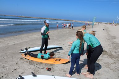 Eine Gruppe von Kindern in Neoprenanzügen beobachtet einen Surflehrer, der Anweisungen am Strand gibt.