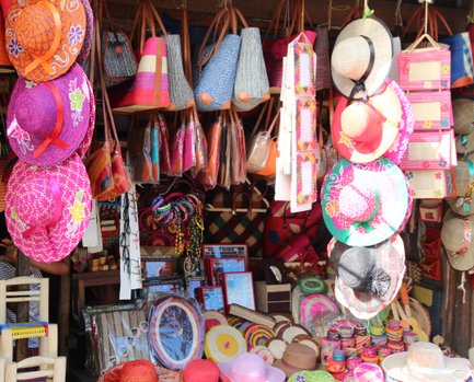 Bunte Strohhüte in verschiedenen Designs hängen an einem Marktstand, umgeben von handgefertigten Taschen und Accessoires.