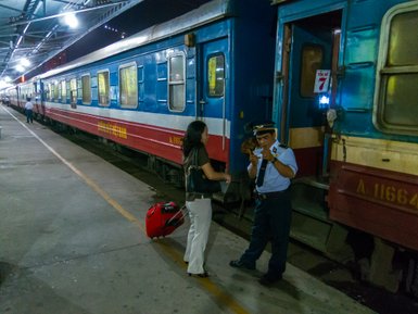 Eine Frau mit einem Koffer spricht mit einem Zugbegleiter an einem Bahnsteig in der Nacht.