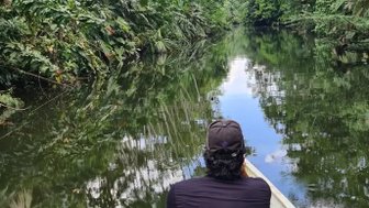 Zwei Personen sitzen in einem Boot auf einem ruhigen Fluss, umgeben von üppigem, grünem Dschungel und spiegelndem Wasser.