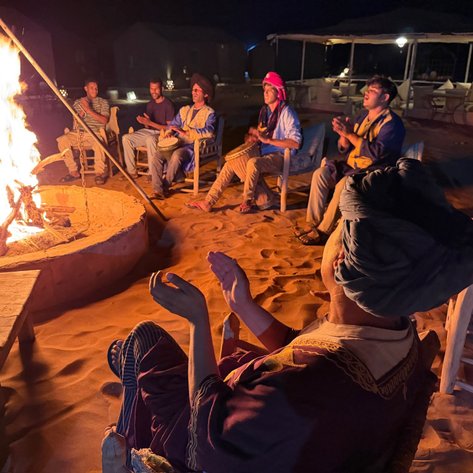 Umgeben von einem lodernden Lagerfeuer sitzen Menschen in einer Wüstenlandschaft und genießen die Nacht.