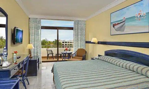 Ein helles Hotelzimmer mit einem großen Bett, einem Schreibtisch und einem Balkon mit Blick auf die tropische Landschaft.