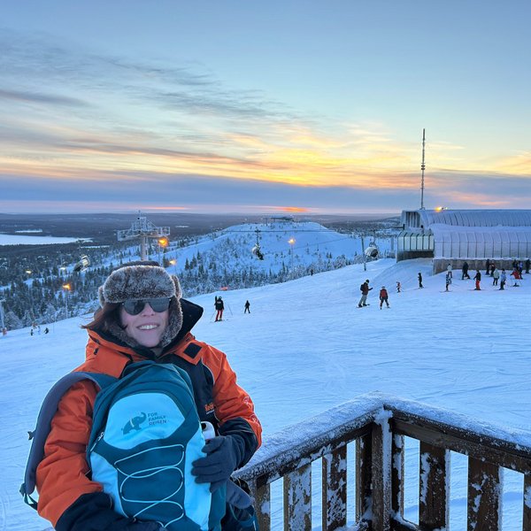 Eine Person mit einer orangefarbenen Jacke und einer blauen Tasche lächelt vor einer verschneiten Skipiste bei Sonnenuntergang.