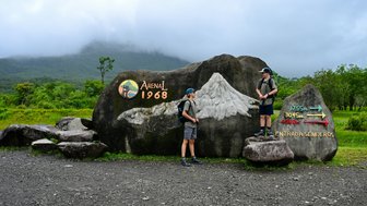 Zwei Wanderer stehen vor einer großen Gesteinsmarkierung im Arenal-Nationalpark, umgeben von üppiger grüner Natur.
