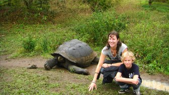 Eine Frau und ein Junge posieren lächelnd neben einer großen Schildkröte in einer grünen, bewaldeten Umgebung.