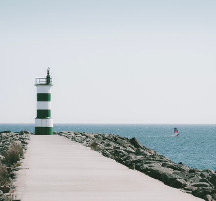 Ein grüner und weißer Leuchtturm steht auf einem Steg, während ein Segelboot sanft über das Wasser gleitet.