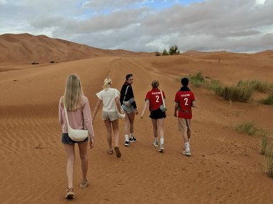 Fünf Jugendliche gehen in einem Sanddünengebiet, umgeben von sanften Hügeln und bewölktem Himmel.