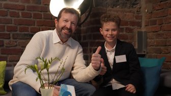 Ein lächelnder Mann und ein Junge sitzen auf einem Sofa, beide zeigen Daumen hoch in einer gemütlichen Umgebung.