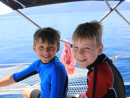 Zwei fröhliche Jungen in Neoprenanzügen sitzen auf einem Boot und genießen die Sonne und das Wasser um sie herum.