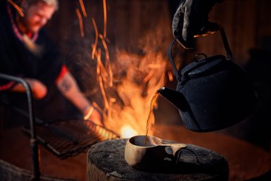Ein warmer Kaffee wird direkt neben dem Feuer aus einer Kanne in eine Tasse gegossen